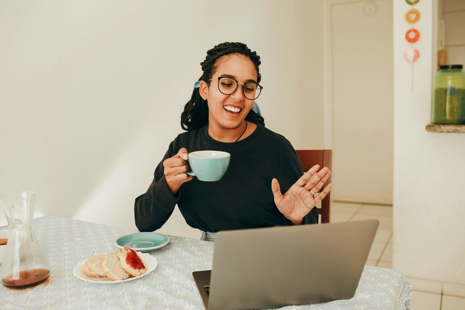 A smiling woman with glasses holds a cup of tea and waves at a video call on her laptop